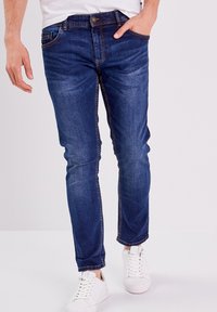 Slim-fit farkut tummansinisellä värillä ja hillityllä pesuefektillä, joissa on viisi taskua ja kontrastiväriset ompeleet. Yhdistetty valkoisiin lenkkareihin.