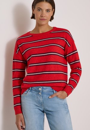 Kvinde iført en rød sweater med sorte og hvide vandrette striber og lyseblå jeans, stående med den ene hånd i lommen mod en ensfarvet væg.