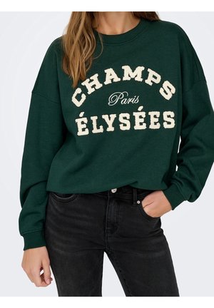 Kvinde iført en mørkegrøn sweatshirt med hvid tekst "Champs Élysées Paris" og sorte jeans, den ene hånd i lommen og beskåret ved skuldrene.
