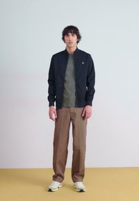 Black lightweight zip-up jacket over a dark green shirt, paired with beige trousers and white sneakers; plain background.