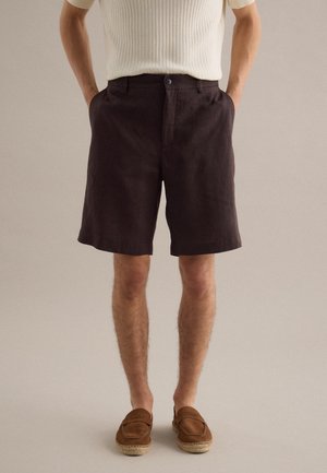 Mann trägt dunkelbraune knielange Shorts, weißes, geripptes Kurzarmshirt und braune Slipper, steht mit den Händen in den Taschen.