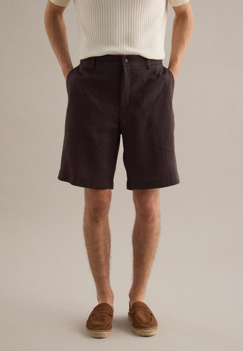 Mann trägt dunkelbraune knielange Shorts, weißes, geripptes Kurzarmshirt und braune Slipper, steht mit den Händen in den Taschen.