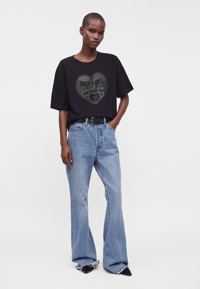 Άτομο που φοράει μαύρο oversized t-shirt με σχέδιο καρδιάς και μπλε τζιν φαρδιάς γραμμής με ξεφτισμένα τελειώματα, στέκεται με το ένα χέρι στην τσέπη.