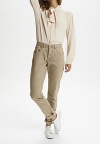 Pantalones beige de cintura alta, con bolsillos frontales y un ajuste esbelto, combinados con una blusa rosa clara que presenta un diseño de cuello con lazo.