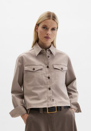 Blonde Frau, die eine beige Jacke mit Knöpfen und Klappentaschen sowie braune Hosen trägt, Hände in den Taschen, neutraler Gesichtsausdruck, einfarbiger Hintergrund.