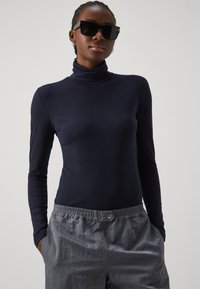 Marineblaues Turtleneck-Oberteil mit langen Ärmeln aus dehnbarem Stoff, kombiniert mit lockeren grauen Shorts mit elastischem Bund und Knopfdetails.