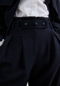 Navy pinstriped tailored trousers with a wide waistband, featuring three dark buttons and a structured fit, made from a smooth fabric.
