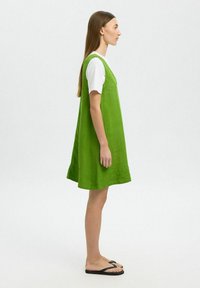 Vestido de lino verde con corte holgado, tirantes en los hombros y dobladillo hasta la rodilla, llevado sobre una camiseta blanca, combinado con chanclas negras.