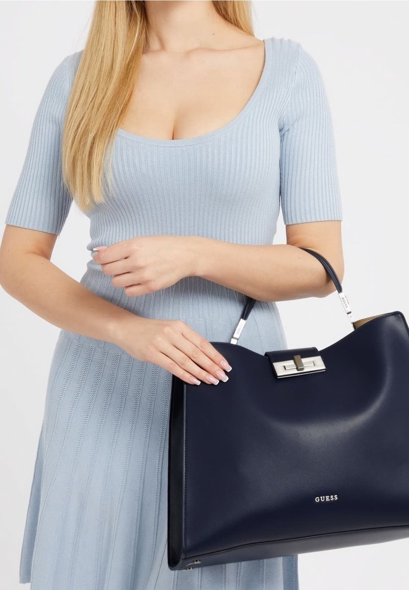 Granatowa skórzana torebka o usztywnionej konstrukcji, z srebrnymi okuciami i górnym uchwytem. Na przedniej części znajduje się widoczne logo marki.