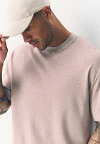 Ljusrosa stickad t-shirt med rund halsringning och korta ärmar. Personen har en beige keps, visar en kortklippt frisyr och tatueringar.