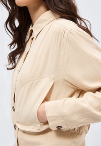 Γυναίκα που φοράει μια μπεζ πουκάμισο με υφή και κουμπιά, με τσέπη στο στήθος, το χέρι μέσα στην τσέπη και με μακριά σγουρά μαλλιά ορατά.