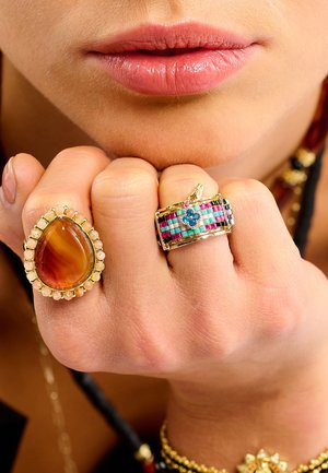 Bagues en or sur les doigts : l'une présente une pierre d'ambre en forme de goutte entourée de petits cristaux ; l'autre affiche des bandes colorées à motifs avec un accent bleu.