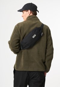 Schwarze Umhängetasche, getragen über einer olivgrünen Fleecejacke, mit geschwungener Form, verstellbarem Riemen und einem kleinen Logopatch an der Seite.