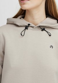 Hoodie beige avec une capuche à cordon. Présente des éléments en métal noir et un petit logo sombre à l'avant. Tissu doux et design décontracté.