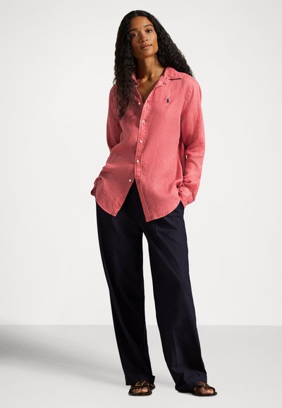 Sieviete ar gariem cirtainiem matiem stāv ar rokām kabatās, valkājot rozā pogājamu kreklu, tumšas platām kājām bikses un brūnus sandales.