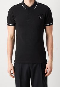 Černé polo tričko vyrobené z bavlny, s žebrovaným límcem s bílým lemem a malým značkovým logem na hrudi. Krátké rukávy.
