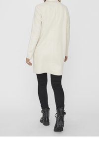 Kermanvalkoinen pitkä takki, jossa on suora leikkaus ja takasaumadetalji, yhdistettynä mustiin leggingseihin ja mustiin nilkkureihin, joissa on soljet.