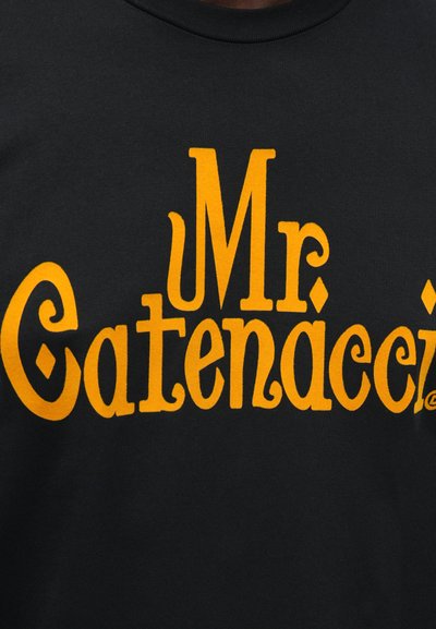 Černé bavlněné tričko s velkým žlutým nápisem "Mr Catenacci" v tučném, stylizovaném písmu s dekorativními prvky.