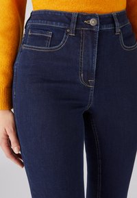 Jeans de mezclilla azul oscuro con un diseño ajustado, ligero desgaste, costuras amarillas y un bolsillo frontal, combinados con un suéter naranja texturizado.