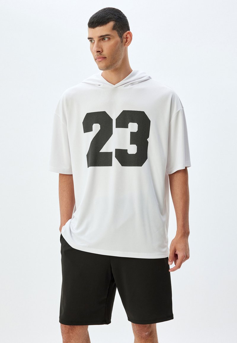Hvid oversized hoodie med korte ærmer, prydet med nummeret "23" i sort mesh-tryk, og parret med sorte atlet shorts, blød stoftekstur.