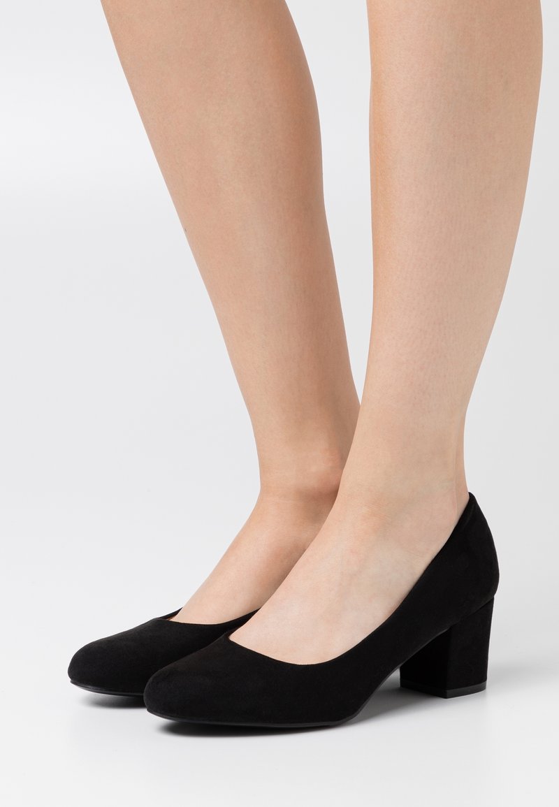 Jambes portant des escarpins en daim noir à talons bas blocs avec bouts arrondis sur fond blanc.
