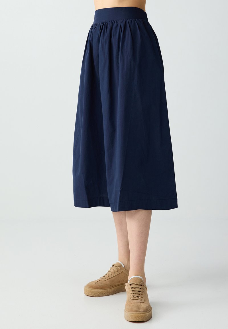 Jupe midi bleu marine avec taille froncée portée avec des baskets beige à lacets sur une personne debout devant un fond uni.