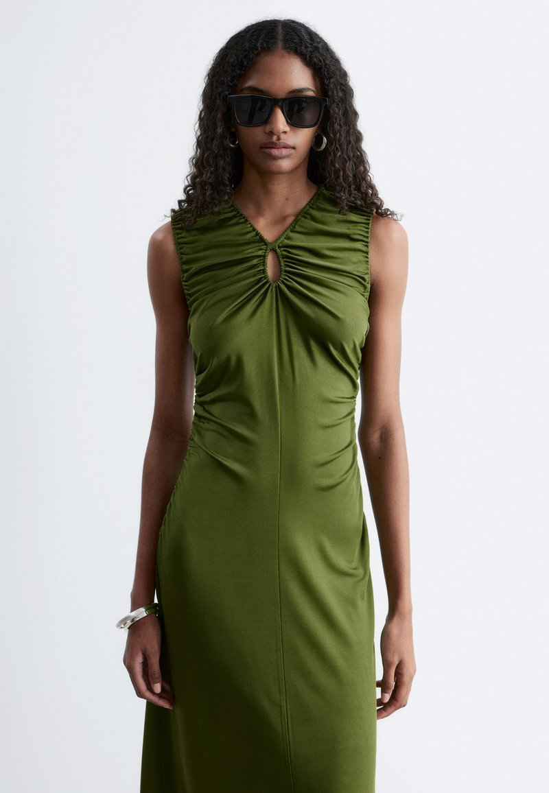 Femme aux longs cheveux bouclés portant des lunettes de soleil noires et une robe olive froncée sans manches sur fond uni.