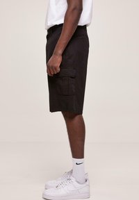 Fekete cargo short, sima textúrájú anyagból készült. Két oldalsó cargo zsebet és egyenes szabást tartalmaz, fehér sportcipővel párosítva.