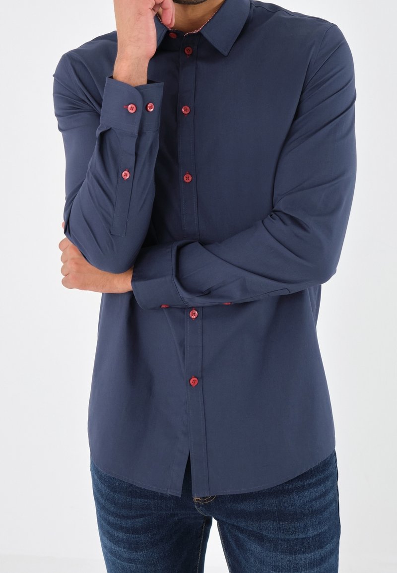Homme portant une chemise bleu marine avec des boutons rouges, debout avec un bras croisé et l'autre main touchant son visage.