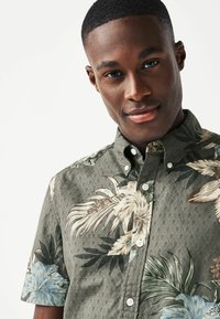 Camisa de botones de manga corta con fondo verde oscuro, adornada con patrones florales multicolores y una superficie texturizada. Diseño de cuello clásico.