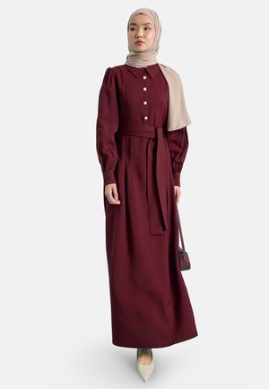 Donna che indossa un lungo vestito bordeaux con bottoni di perle, hijab beige, scarpe bianche a punta e che porta una piccola borsa bordeaux.