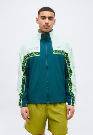 Mladý muž nosiaci tyrkysovobielu bundu na zips so zelenými pruhmi s vzorom a žlté šortky, stojaci pred jednoduchým pozadím.