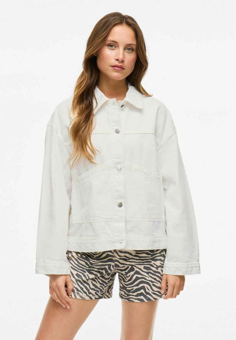 Chaqueta de mezclilla blanca con cierre de botones, grandes bolsillos y un corte relajado, combinada con pantalones cortos beige con un patrón de rayas de cebra.