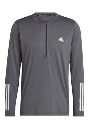 Personne portant un t-shirt gris à manches longues Adidas et un pantalon noir, tenant un kettlebell de 8 kg dans une main, debout en position écartée.