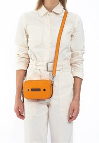 Oranje textuur leren schoudertas met een rechthoekige vorm, met zwarte accenten en een ritsluiting, gedragen met een lichtgekleurde jumpsuit.