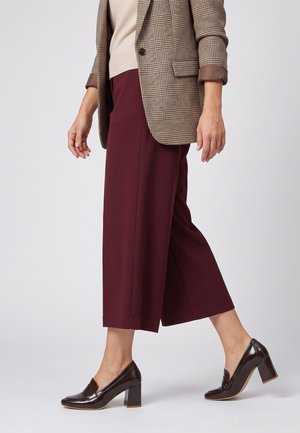 Person iført bordeaux bukser med vide ben, en beige sweater, en ternet blazer og mørkebrune sko med blokhæl, gående mod en hvid baggrund.