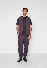 Herrset i två delar med ärmlös lila väst med röda detaljer och logotyper, kombinerad med matchande lila byxor och svarta sneakers.