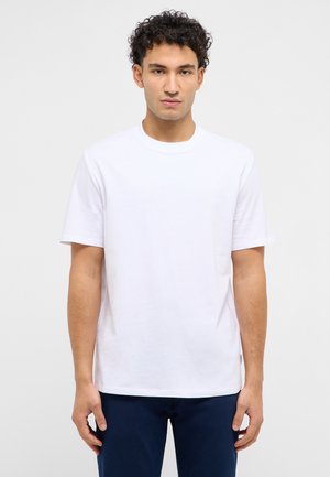 Junger Mann mit kurzen lockigen Haaren, der ein einfaches weißes kurzärmliges T-Shirt und dunkelblaue Hose trägt und vor einem weißen Hintergrund steht.