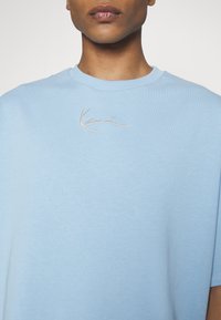 Heledad sinistee lühikese varrukaga T-särk, mis on valmistatud pehmest puuvillast, featuring kaarjas kaelus ja valge tikandiga "Kanye" logo.