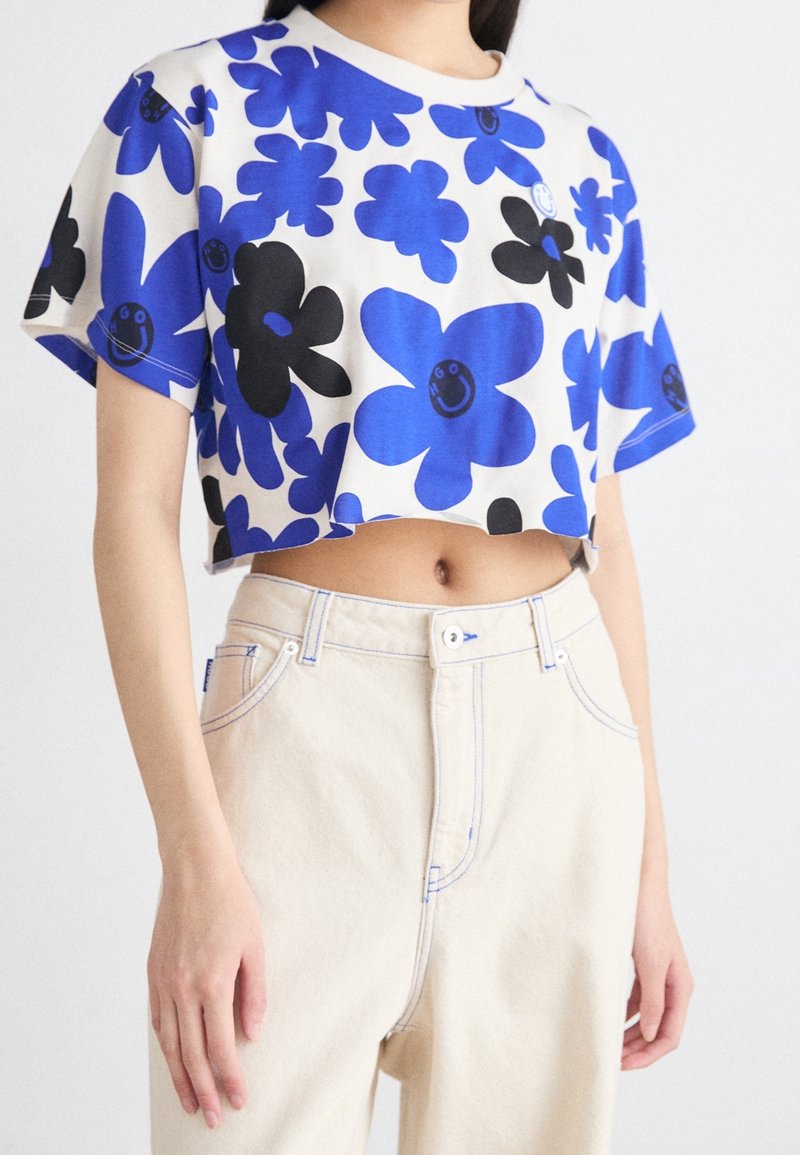 Camiseta corta de flores azules con detalles en negro sobre un fondo claro, mangas cortas y cuello redondo; presenta flores sonrientes y juguetonas.