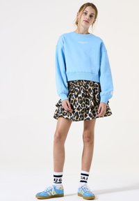 Sudadera recortada de color azul claro con texto blanco, combinada con una falda de leopardo con volantes. Acentuada con zapatillas deportivas azules y calcetines a rayas.