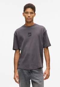 Tamsiai pilkas trumpas rankovėmis marškinėliai su tekstūruotu, vaflinio rašto dizainu ir kvadratiniu logotipo lipduku ant krūtinės. Kasdienis fasonas ir apvalus iškirpimas.