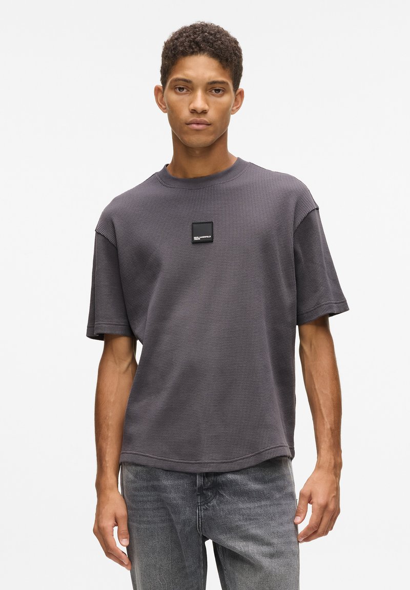 Tamsiai pilkas trumpas rankovėmis marškinėliai su tekstūruotu, vaflinio rašto dizainu ir kvadratiniu logotipo lipduku ant krūtinės. Kasdienis fasonas ir apvalus iškirpimas.