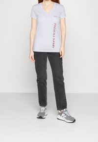 Camiseta blanca de cuello en V con gráfico rojo de "Armani Exchange" a lo largo del lado, combinada con jeans gris oscuro y zapatillas gris claro.