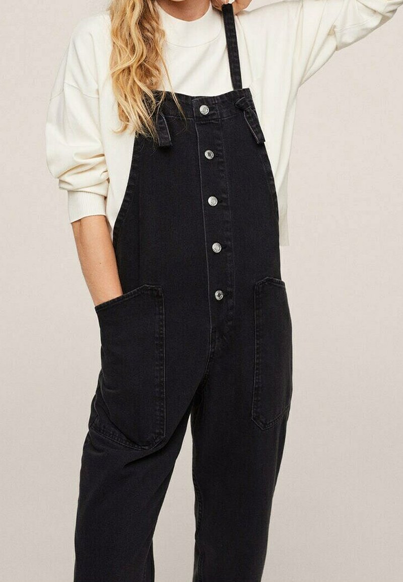 Femme portant une salopette en denim noir boutonnée avec de grandes poches avant et une chemise blanche à manches longues, main dans la poche, sur fond neutre.
