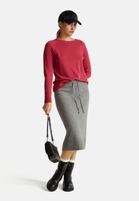 Czerwony sweter z dzianiny, szara spódnica midi z prążkowanego materiału z troczkiem, czarne buty za kostkę na sznurówki oraz czarna czapka. Modelka trzyma małą czarną torebkę.