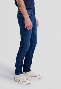 Bărbat purtând blugi skinny albastru închis și adidași înalți albi din pânză, stând pe un fundal gri deschis.