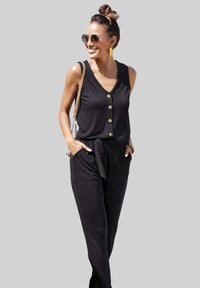 Un mono negro sin mangas con botón al frente, cintura ajustada, bolsillos laterales y un corte relajado, complementado con unos pendientes amarillos y gafas de sol.