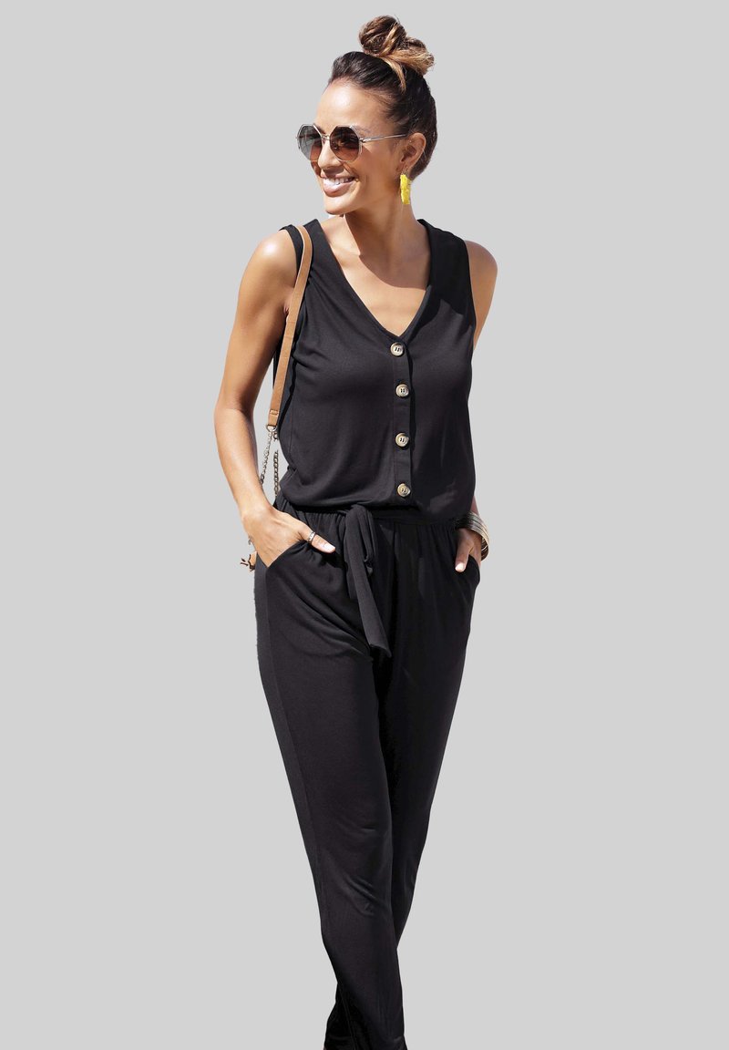 Un mono negro sin mangas con botón al frente, cintura ajustada, bolsillos laterales y un corte relajado, complementado con unos pendientes amarillos y gafas de sol.