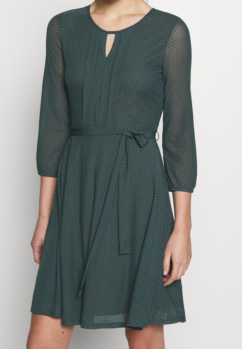 Robe vert foncé longueur genou avec pois noirs, manches 3/4 transparentes, encolure ronde avec petite découpe à l'avant et ceinture nouée à la taille.